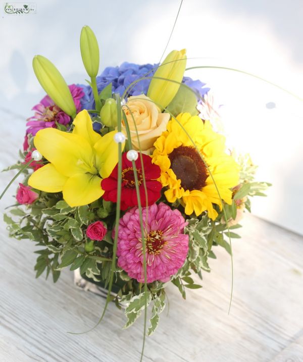 Rainbow colored table decoration in a glass cube (hydrangea, sunflower, rose, lily)